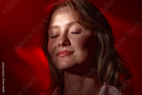 Freckled young woman with closed eyes enjoying a peaceful, relaxed moment bathed in warm red light. The intimate portrait emphasizes natural skin texture, calm expression, and soft cinematic lighting.