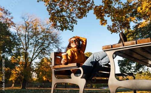 Relaxed woman sitting on bench with feet up and takeaway coffee during sunny autumn day in park