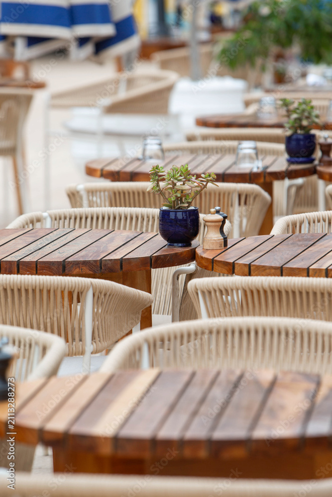 Fototapeta premium Cozy outdoor seating area with wooden tables, wicker chairs, and small potted plants at a hotel restaurant near a swimming pool. Elegant resort dining atmosphere with shallow depth of field