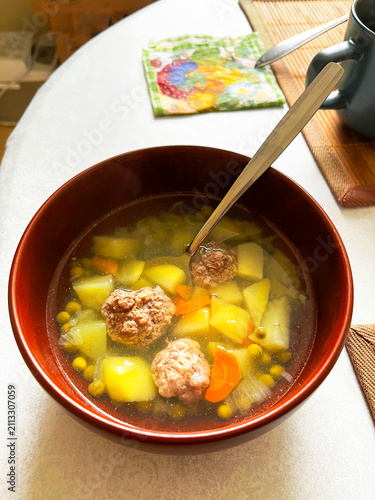 A Ceramic Bowl of Soup with Meatballs, Green Peas and other Vegetables on a Table covered with a White Tablecloth. Traditional Slavic Dish. Homemade Food.
