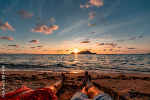 Couple relaxing on a sand beach while watching the sunset, legs visible, holiday concept