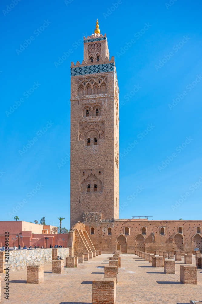 Obraz premium Kutubiyya Mosque stands tall against a clear blue sky in Marrakesh. Visitors walk in the area, taking in the structure and its surroundings during the day.