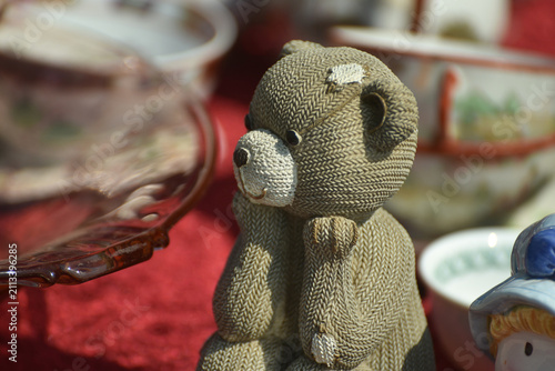 Old teddy bear sitting with its paws under the chin among vintage objects in a flea market, creating a nostalgic atmosphere of childhood memories and second hand treasures.
