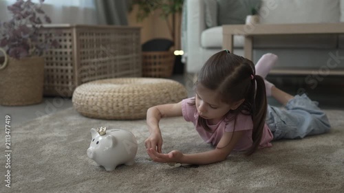 Little girl putting coins into piggy bank lying on carpet