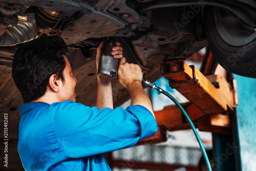 Car Repair Performed with a Pneumatic Impact Wrench as an Auto Mechanic Works Beneath a Raised Vehicle Inside an Automotive Service Garage During Routine Maintenance