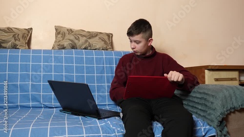 Teenager is sitting on a couch, continuously working with a tablet and laptop, checking, copying or processing study materials.