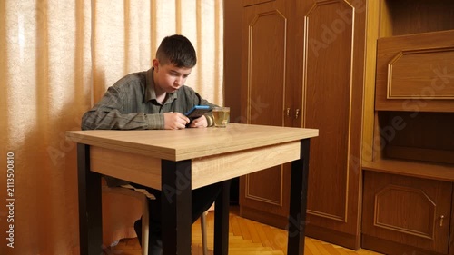 Teenager is sitting at a table, actively playing on a handheld game console while occasionally drinking tea from a clear glass.