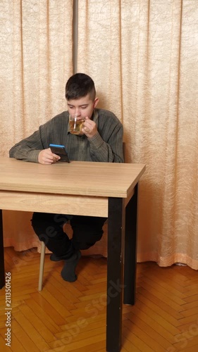 Teenager is sitting at a table, actively playing on a handheld game console while slowly drinking hot tea from a glass.