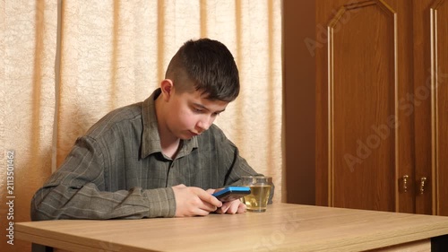 Teenager is sitting at a table, actively playing on a handheld game console, carefully controlling the game with a glass of hot tea nearby.