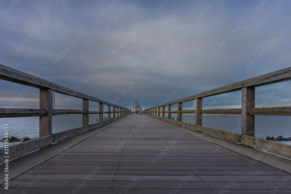 Obraz premium Low angle perspective of wooden pier boardwalk extending into calm ocean under clouds