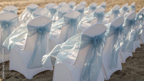 Rows of white chairs with transparent umbrellas and blue bows arranged on the sand. Outdoor wedding setup. Elegant and decorative event seating arrangement.