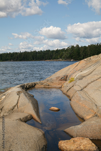 Sunlit granite rock pool with scattered boulders opening onto a calm forested lake.