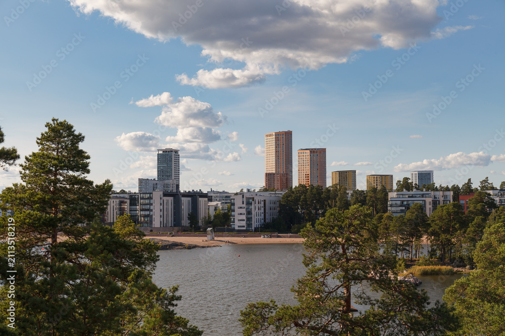 Obraz premium High-rise skyline of Helsinki in Finland viewed across a calm bay lined with pine trees and a sandy beach.