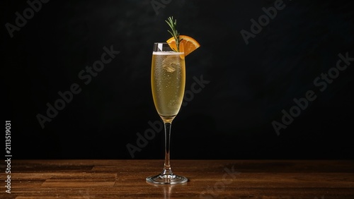 A glass of champagne or sparkling wine with an orange garnish on a dark background.