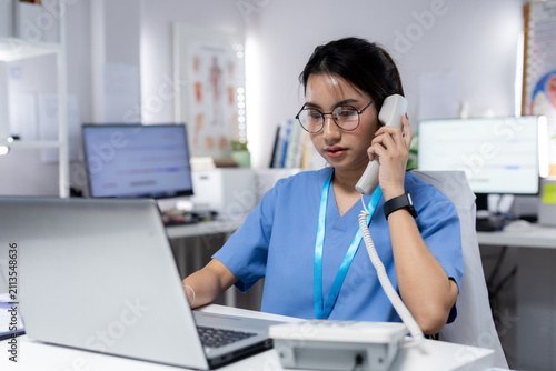 professional answering patient phone call while working on laptop in clinic, representing medical communication, appointment scheduling, healthcare support, patient services, clinical administration.