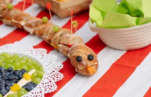 Картината върху платно Creative caterpillar shaped baguette sandwich with cheese, vegetables and blueberries served on a striped picnic tablecloth