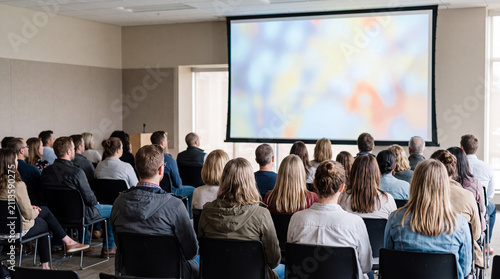 Audience seated in rows watching a large projection screen in a dim overflow room, seen from behind