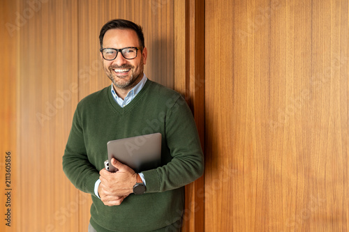 Businessman in eyeglasses holding a digital tablet, smiling and looking confident, embodying professionalism, leadership, and success in a modern office environment