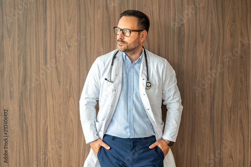 Confident medical practitioner wearing eyeglasses and lab coat, looking away, reflecting on healthcare challenges and professional dedication while standing against a textured backdrop