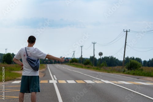 Man with a bag hitchhikes on a rural road by extending his thumb. The wide road stretches into the distance with a vehicle approaching, highlighting travel and adventure themes.