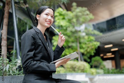 Smiling businesswoman holds a digital tablet and stylus in a modern green office courtyard, representing professional confidence, career planning, mobile work, leadership, and corporate success.