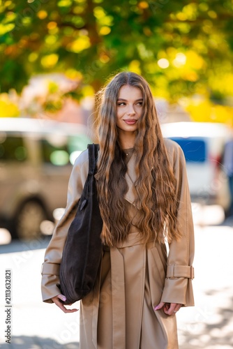 Young woman with long wavy brown hair in tan trench coat and high waist skirt carrying shoulder bag outdoors in soft sunlight with warm blurred greenery behind, with woman, portrait, hair, longhair