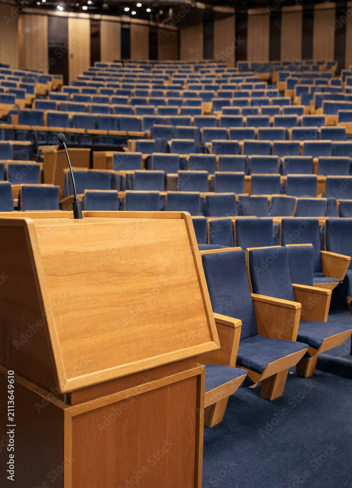 Fototapeta premium Wooden podium with microphone stands in front of rows of blue upholstered seats in an empty auditorium. The space conveys readiness for a lecture or presentation.