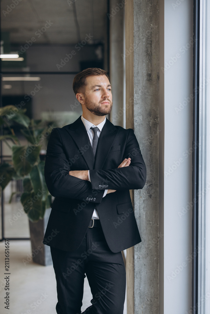 Fototapeta premium Confident businessman in suit with folded arms standing by an office window looking into the future