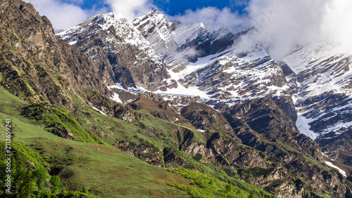Himachal Pradesh snow capped Himalayan mountain peaks rising above green grassy valley slopes under blue sky