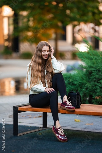 Young woman sitting on park bench in autumn glow with long wavy hair, casual jacket, jeans, burgundy sneakers beside handbag, lush greenery and blurred city backdrop
