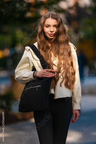 Portrait of young woman outdoors with long wavy hair, cream jacket, black tote bag, soft smile, and warm sunlit urban park setting with blurred background, with woman, portrait, outdoor, lighting