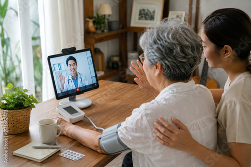 Elderly Asian woman and daughter having telemedicine consultation with doctor on tablet at home