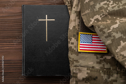 Christian cross on black book beside military camouflage uniform with American flag US, concept of faith and military service