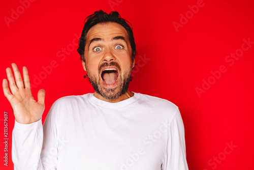 Studio portrait of a surprised shocked young caucasian man with dark hair and beard wearing white long sleeve t shirt wide eyes open mouth waving hand gesturing greeting or shock on red background