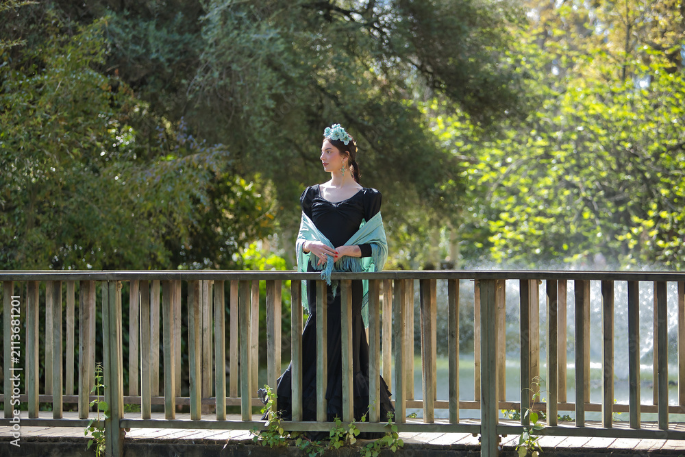 Fototapeta premium A young woman from Andalusia wearing a black flamenco dress with ruffles and an aquamarine shawl, with flowers in her hair, posing on a wooden bridge in an outdoor park. Concept: dance, art.