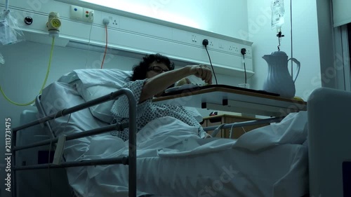 Woman eating sandwich in hospital bed, curly dark hair lady is having her breakfast or lunch from tray in a dim lit room, low angle view.