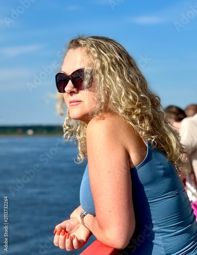 Woman with blonde curly hair in sunglasses stands on a boat and looks at the water on a sunny summer day
