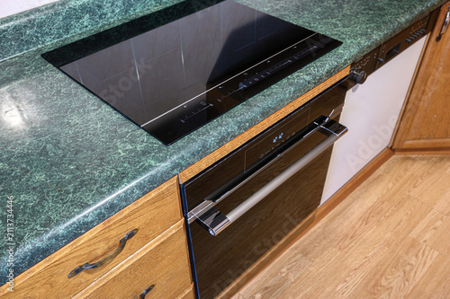 kitchen features a green marble countertop with a speckled pattern, a built‑in black induction cooktop, a black dishwasher with a digital display, and wooden cabinets with dark metal handles