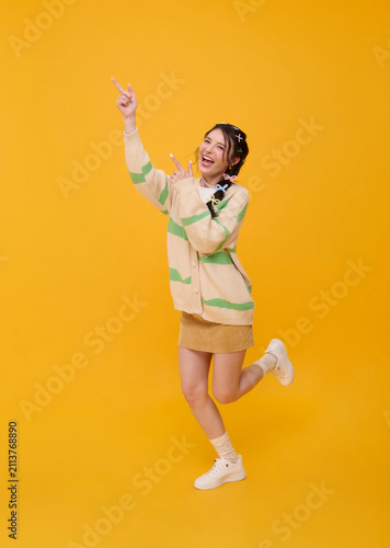 Asian woman pointing upward with joyful expression isolated on yellow studio background promoting positive lifestyle new ideas success celebration advertising and youthful confidence.