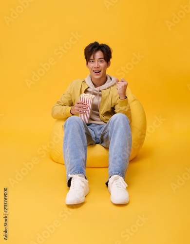 Movie entertainment concept with excited Asian man holding popcorn and cheering on beanbag in studio expressing leisure streaming enjoyment positive emotion and youthful home cinema lifestyle.
