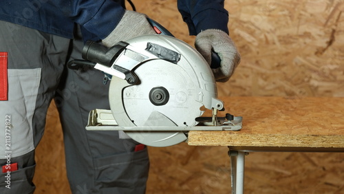 Builder saws a board with a circular saw cutting a wooden plank in a joinery. Carpenter uses portable circular saw tool to cut wood at wooden table in workshop. Worker sawing pine board. Real image
