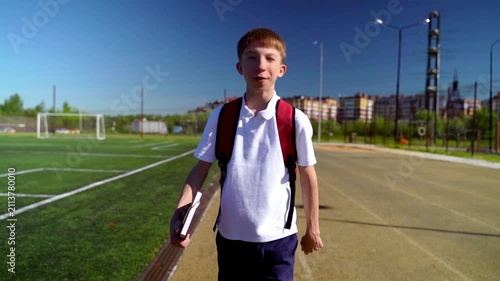 Young boy walks toward camera on sunny day. He carries backpack and school book. He smiles slightly, looking calm and cheerful. Sidewalk runs beside green soccer field