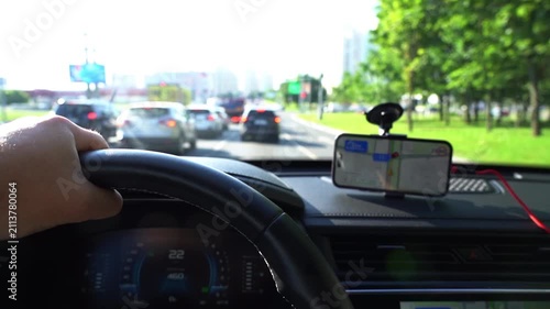 Driver grips steering wheel, focused on road ahead. Phone mounted, displaying navigation map. Cars move slowly through urban traffic. Scene suggests commuting or navigation during rush hour