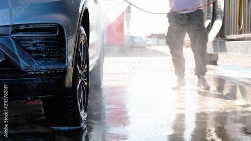 Person washes car with high-pressure hose. Water sprays onto vehicle's dark, wet surface. Ground reflects light, showing wetness and motion. Worker's posture suggests focus on cleaning