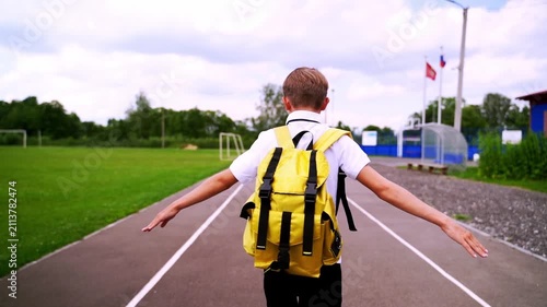 Child stands on track with open arms. Yellow backpack suggests adventure or new beginnings. Background shows green fields and distant flags. Feeling of freedom and excitement is clear