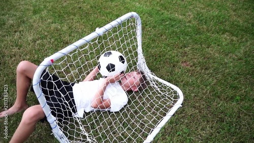Child sleeps with soccer ball inside goal frame. Grassy field surrounds playful resting scene. Child smiles peacefully, resting on net. Footage perfect for sports, child, or leisure themes