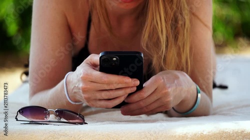 Woman relaxes on beach towel, scrolling phone. Sunglasses rest beside her, out of use. Soft sunlight bathes her bare arms and skin. She holds phone steadily, focused on screen. Calm, leisurely mood