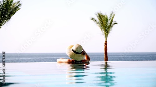 Woman relaxes in infinity pool beside ocean. Palm trees frame tranquil seaside scene. Soft light enhances peaceful vacation atmosphere. Ideal for luxury travel or wellness campaigns