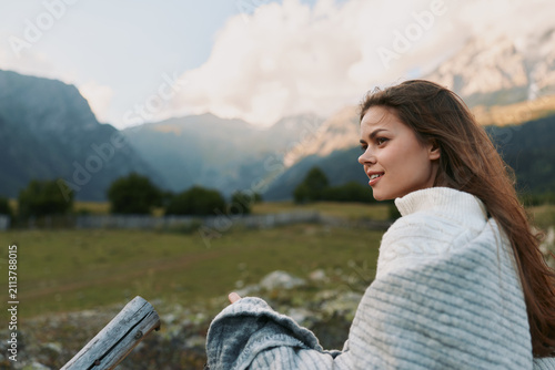 Foto Young woman wrapped in a white shawl sits beside a wooden fence, smiling softly as majestic mountains rise in the distance on a bright, clear day