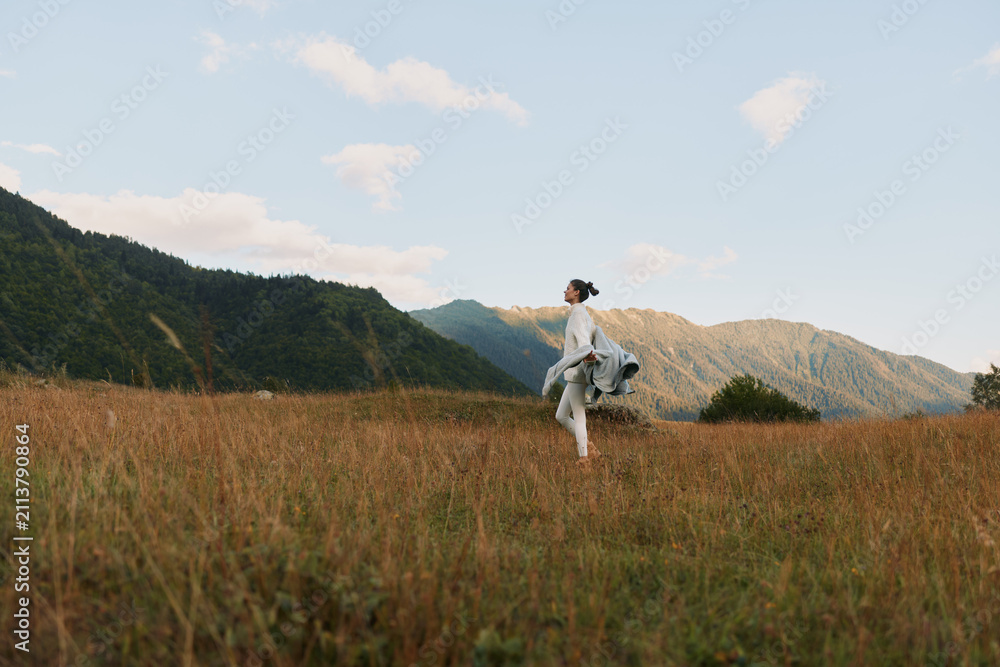 Obraz premium Woman walking in a grassy field near mountains under a blue sky with clouds. Lifestyle concept with nature, freedom, and outdoor activity in soft natural light.